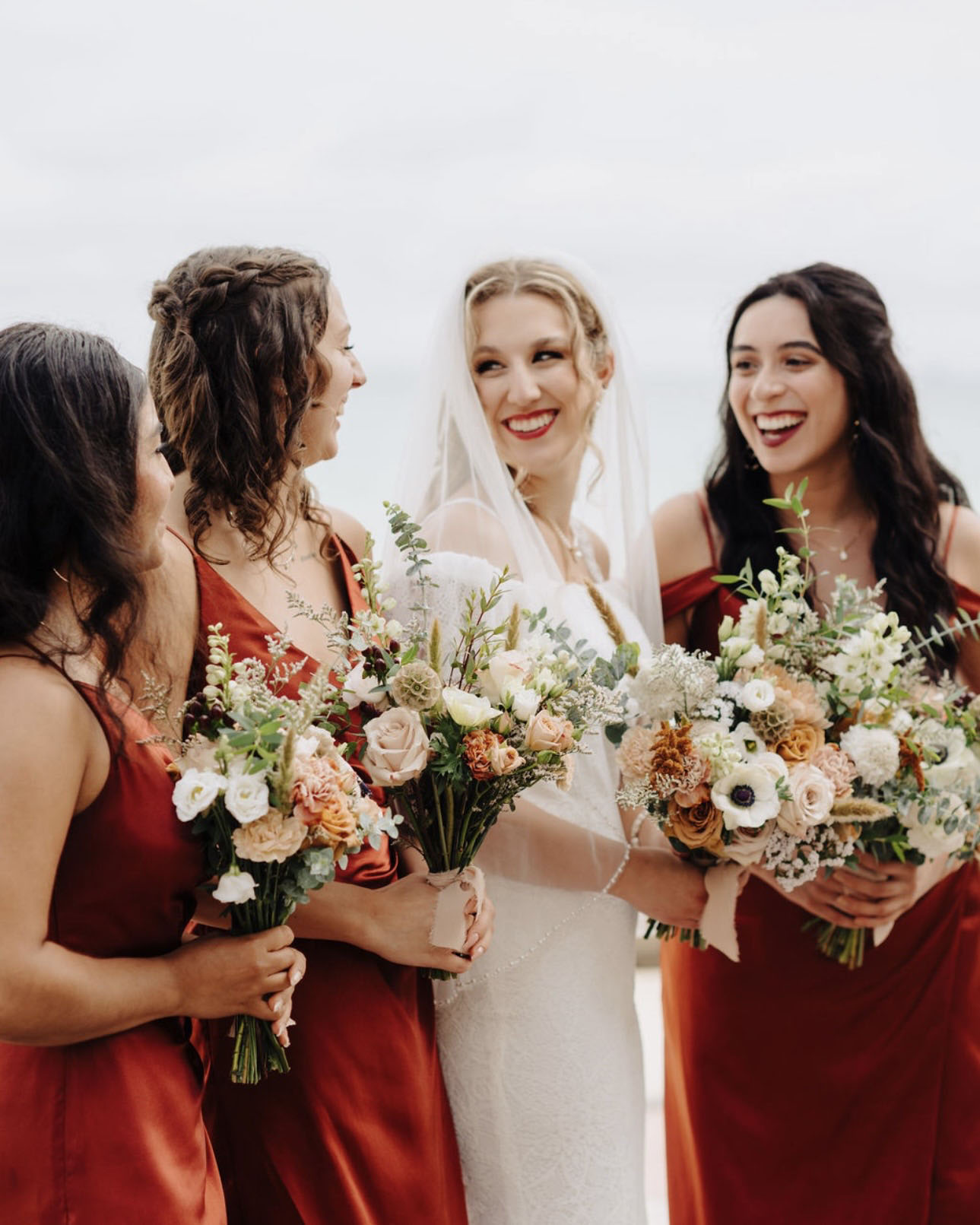 A bride in a white veil laughing with three bridesmaids wearing rust-colored satin dresses and holding wildflower bouquets.