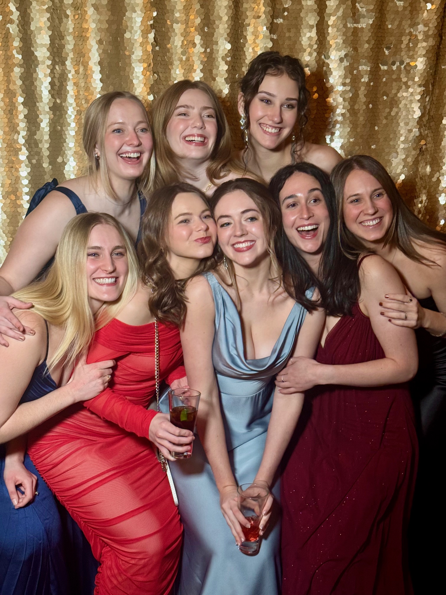 A group of seven smiling women in formal evening gowns posing in front of a shimmering gold sequin photo booth backdrop at a gala event.